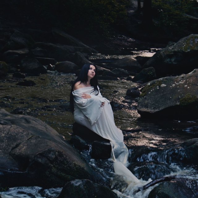 Pochette de Under The Same Moon : une femme en robe blanche assise sur des rochers au bord d’un ruisseau