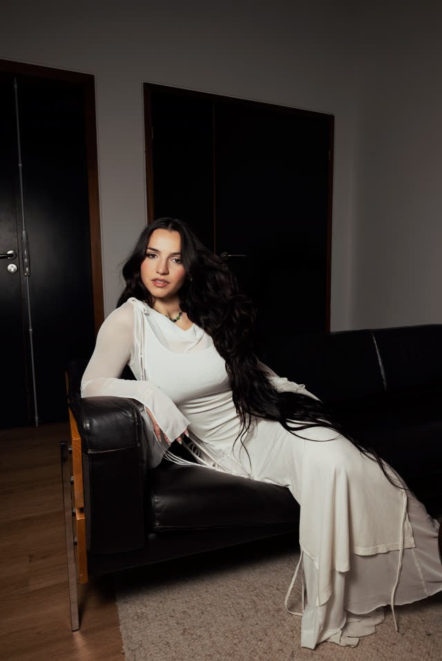Myla en robe blanche, assise sur un canapé en cuir noir