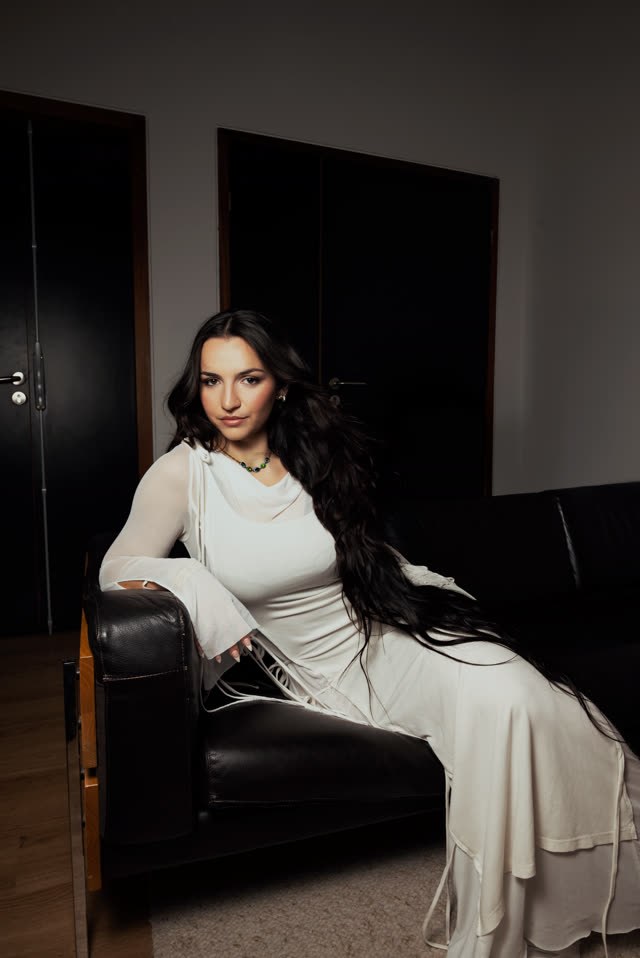 Myla en robe blanche, allongée sur un canapé en cuir noir