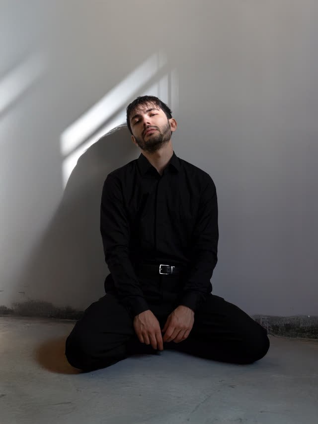 Rosenfeld en noir, à genoux contre un mur blanc, dans la lumière d’une fenêtre