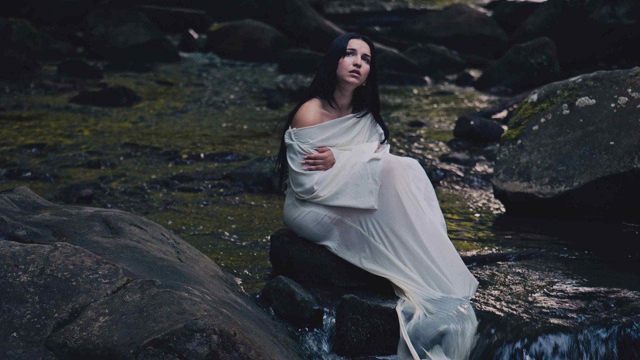 Une scène du clip de Under The Same Moon : Myla en robe blanche, assise sur des rochers au bord d’un ruisseau