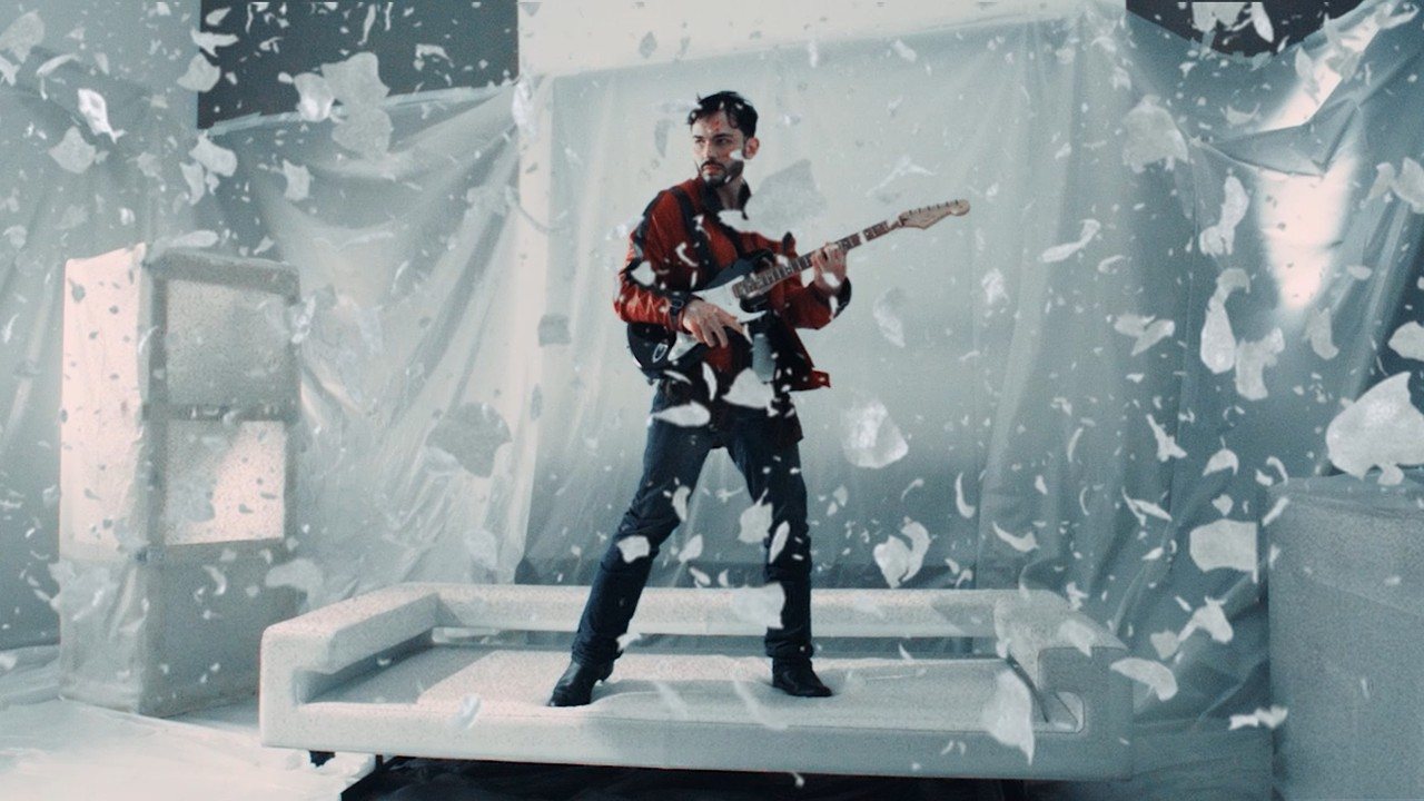 Rosenfeld joue de la guitare électrique dans un décor blanc, entouré de fragments qui volent